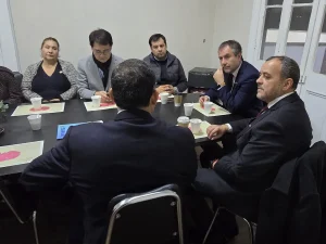 El gerente general de Sercotec encabeza una reunión de trabajo junto a representantes del barrio Patronato y ejecutivos de Sercotec sentados alrededor de una mesa larga y negra. En el centro de la mesa hay una planta con flores rosadas, vasos y folletos, mientras los asistentes escuchan con atención.