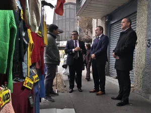 El gerente general de Sercotec, junto al director regional de Sercotec y el presidente de la Cámara de Comercio de Patronato conversan en la acera con un comerciante frente a un local de ropa expuesta al aire libre con carteles amarillos de ofertas. Al fondo se ve la cortina metálica cerrada de otra tienda.
