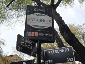 Primer plano de un poste con carteles informativos en la vía pública. El letrero superior promociona el "Barrio Patronato" como barrio comercial y multicultural, mientras que los letreros inferiores indican los cruces de las calles Sagrado Corazón y Patronato con sus respectivas numeraciones.