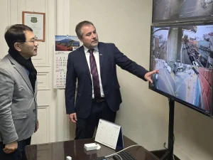 El gerente general de Sercotec y el presidente de la Asociación Coreana en Chile en una oficina observan una pantalla de televisión que muestra múltiples transmisiones en tiempo real de cámaras de seguridad de la calle. El gerente general de Sercotec apunta con su mano hacia uno de los cuadrantes del monitor.