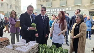 En una feria al aire libre con edificios urbanos al fondo, el gerente general de Sercotec observa sonriente una alcachofa fresca que sostiene en su mano. A su lado, el ministro de Economía, junto a mujeres representantes de las ferias libres—una de ellas con delantal blanco de cocina—, observan la escena de manera amigable. En el primer plano se aprecian múltiples bandejas apiladas con huevos blancos y una hilera de alcachofas frescas sobre el mostrador.