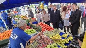 Vista en perspectiva elevada de un colorido puesto de feria. En la parte inferior, un feriante de espaldas con una camiseta azul que dice "MÁS Y MEJORES FERIAS LIBRES PARA CHILE!" estira su brazo para mostrar una fresa a un grupo de personas. Entre los visitantes destacan el ministro de Economía y el gerente general de Sercotec que sonríen mientras recorren el lugar. El puesto rebosa de frutas frescas organizadas: plátanos amarillos, manzanas verdes, uvas, fresas envasadas, piñas y tomates rojos. Al fondo se observa el movimiento de clientes caminando por la feria.