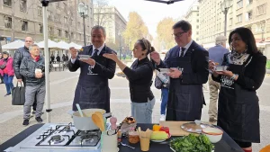 El gerente general de Sercotec y el ministro de Economía, vestidos con delantales negros de cocina sobre su ropa, sostienen pequeños recipientes con comida lista para degustar en medio de una plaza pública. Una chef con micrófono de cintillo les ofrece una muestra desde un mesón de cocina portátil equipado con una encimera a gas, ollas, verduras frescas (como espinacas y pimientos) y utensilios. De fondo se aprecian transeúntes, árboles con tonos otoñales y la arquitectura de una avenida céntrica.