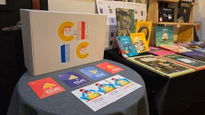 Primer plano de una mesa con productos de la cooperativa Clic, incluyendo una caja grande con ilustraciones, libros infantiles coloridos y postales promocionales.