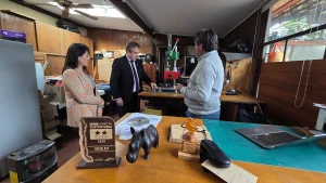Una vista amplia del taller interior de la empresa Reciklast. En primer plano, una mesa de madera muestra el premio "Sello Impacta Sustentable 2025", junto a otros elementos decorativos. Al fondo, tres personas conversan cerca de una máquina.