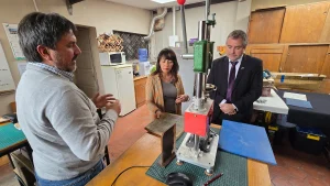 El gerente general de Sercotec y la jefa de Sostenibilidad FEN UChile escuchan atentamente la explicación del fundador de Reciklast, quien gesticula con las manos. Están parados junto a una prensa mecánica en el taller.