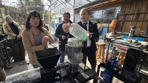 El gerente general de Sercotec vierte virutas de plástico verde de una botella de plástico grande en el embudo de la máquina de reciclaje, mientras la jefa de Sostenibilidad FEN UChile señala la máquina y el fundador de Reciklast observa.