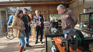La jefa de Sostenibilidad FEN UChile, el fundador de Reciklast y el gerente general de Sercotec observan a un empleado con una sudadera con capucha operar una máquina de extrusión de filamentos de plástico en el taller exterior.