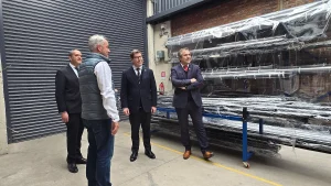 El director regional de Sercotec, el fundador de Garage de Proyectos, el ministro de Economía y el gerente general de Sercotec conversan de pie en un pasillo de la fábrica, junto a una estantería con perfiles metálicos envueltos en plástico.