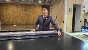 El gerente general de Sercotec, vistiendo traje y corbata roja, manipula y extiende un rollo de plástico transparente sobre una mesa de trabajo negra en el taller.