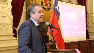 El gerente general de Sercotec sonríe mientras habla por un micrófono junto a la bandera chilena y una pantalla de proyección iluminada.