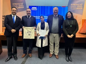 Cinco personas posan para una foto sobre un escenario. Al centro, una representante de las cooperativas sostiene un documento y el gerente general de Sercotec una bolsa de tela. Están flanqueados por el seremi de Economía, el director regional de Sercotec y una representante de las cooperativas beneficiarias quien posa sonriente con collar verde.