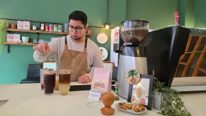 Primer plano de uno de los dueños de Inti Mapu, usando delantal y lentes, preparando bebidas detrás del mostrador. Hay un molinillo de café y estantes con productos de fondo.